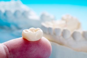 In the foreground is a dental crown resting on a fingertip with a blurry model of a jaw in the background